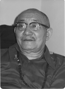 Portrait of an elderly man wearing glasses and a beaded necklace.